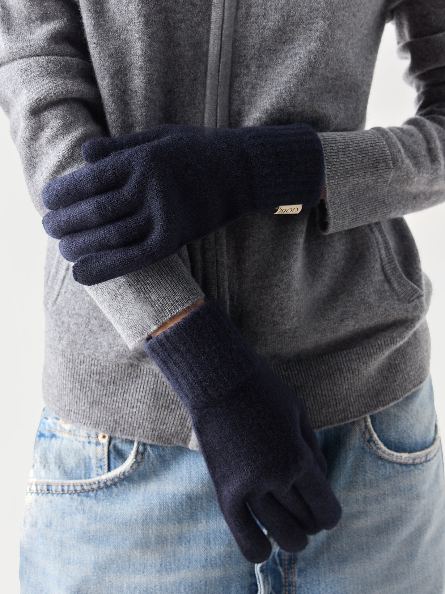 Person wearing navy gloves with a gray jacket and blue jeans on a light background